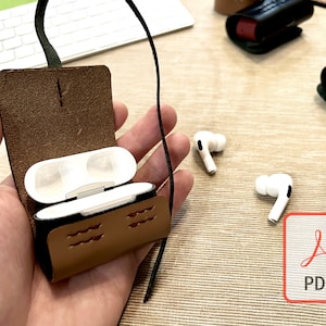 May include: A brown leather case with a flap that opens to reveal a white wireless earbud charging case. The case has a black strap attached to it. Two white wireless earbuds are lying on a brown surface.