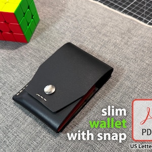 May include: A black leather slim wallet with a snap closure. The wallet has a minimalist design with white stitching and a silver snap. The text "slim wallet with snap" is visible in the image.