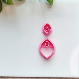 May include: Two pink polymer clay cutters in an abstract leaf shape. The cutters are in two sizes, one smaller and one larger. The cutters are on a white surface.