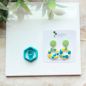 May include: A pair of handmade polymer clay earrings with a green stud and a colorful, patterned hoop design. The earrings feature yellow, blue, and white accents. A turquoise hexagon-shaped clay cutter is also visible.