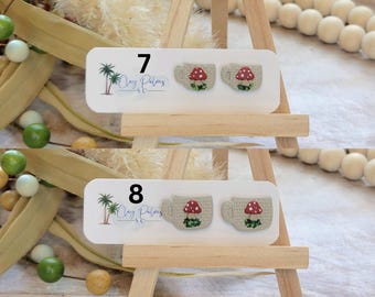 May include: Two pairs of mushroom-themed earrings displayed on a wooden easel. Each earring is shaped like a teacup with a red mushroom design. The earrings are on a white card with the text "7" and "8" and the logo "Clay Palms SC".