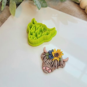 May include: A green plastic cookie cutter shaped like a Highland cow's head. A brown and white fondant Highland cow head with a yellow sunflower and blue flowers on top is next to the cutter.