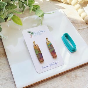 May include: A pair of handmade earrings with a colorful geometric design, displayed on a white card with the text "Clay Palms". A turquoise oval-shaped cutter is next to the earrings. The earrings have gold-colored posts.