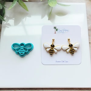 May include: A teal bee-shaped clay cutter next to a pair of bee-shaped earrings. The earrings have white wings, gold accents, and black and yellow striped bodies. The earrings are on a white card with text.