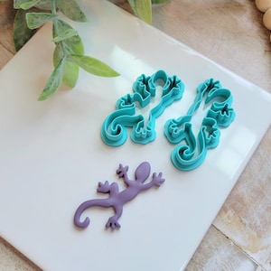 May include: A purple lizard figurine and two teal lizard-shaped cookie cutters rest on a white tile. The cookie cutters have a detailed, open design. The lizard figurine is a solid color and has outstretched legs.