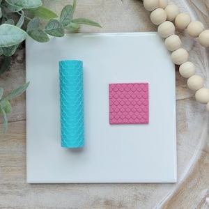 May include: Two silicone rolling pins with a mermaid scale pattern. One is a long cylinder and the other is a square. The cylinder is turquoise blue and the square is pink.
