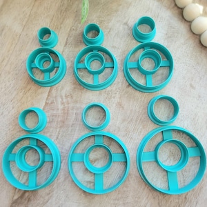 May include: Set of six turquoise plastic cookie cutters with different sizes and shapes. The cutters feature a ring with a smaller circle in the center and four spokes extending from the center circle to the outer ring.