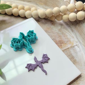 May include: Two teal dragon-shaped cookie cutters and a small purple dragon cookie are displayed on a white surface. The cutters are made of plastic and have detailed wing and body designs. The cookie is a finished product, also with detailed wings.