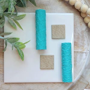 May include: A collection of teal and tan textured clay pieces arranged on a white square tile. The teal pieces are cylindrical, while the tan pieces are square. The arrangement is set against a wooden background with greenery and wooden beads.