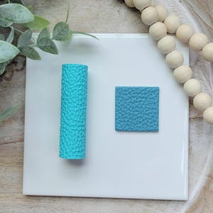 May include: A turquoise textured cylinder and a square of matching material sit on a white surface. The cylinder is upright, and the square is to its right. Decorative wooden beads and greenery are in the background.