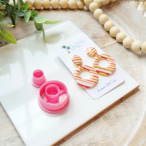 May include: A pair of handmade, striped hoop earrings in red, yellow, and white, resting on a white tile. A pink clay cutter and a smaller pink cutter are also present. The earrings are on a card that reads "Handmade & Style Culture."
