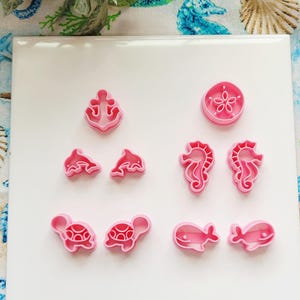 May include: Pink plastic cookie cutters in various sea creature shapes, including an anchor, dolphins, seahorses, turtles, and whales. The cutters are arranged on a white surface, perfect for making ocean-themed cookies.
