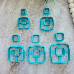 May include: Set of turquoise blue plastic square cookie cutters of various sizes. The cutters are arranged in a pattern on a wooden surface.