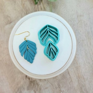 May include: A pair of teal blue leaf-shaped earrings with a gold hook. The earrings are displayed on a white circular plate with two teal blue leaf-shaped cookie cutters.