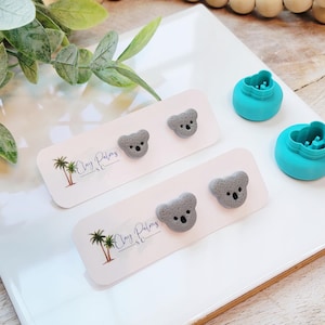 May include: Gray koala bear stud earrings on white cards with the text "Clay Palms SC". Two turquoise clay cutters are also visible. The earrings and cutters are on a white surface with greenery.