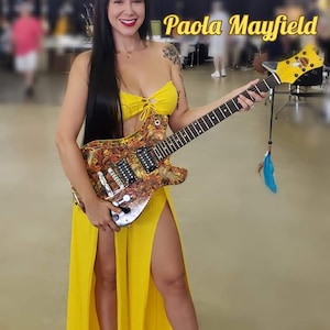Puede incluir: Una mujer con un vestido amarillo y un top amarillo sostiene una guitarra eléctrica personalizada con un diseño abstracto colorido. La guitarra tiene una placa de protección negra y una pala dorada. La mujer está de pie en una habitación con un suelo de color claro. El texto "Paola Mayfield" es visible en la imagen.