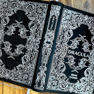 K&ouml;nnte beinhalten: Ein schwarzes Hardcover-Buch mit einem wei&szlig;en, verzierten Design auf dem Einband. Der Titel "Dracula" ist in Wei&szlig; auf dem vorderen Einband gedruckt. Der Autor "Bram Stoker" ist in Wei&szlig; auf der Unterseite des vorderen Einbands gedruckt.