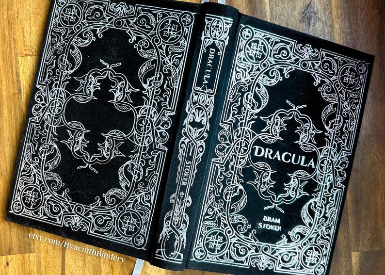 K&ouml;nnte beinhalten: Ein schwarzes Hardcover-Buch mit einem wei&szlig;en, verzierten Design auf dem Einband. Der Titel "Dracula" ist in Wei&szlig; auf dem vorderen Einband gedruckt. Der Autor "Bram Stoker" ist in Wei&szlig; auf der Unterseite des vorderen Einbands gedruckt.