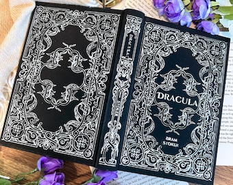 Puede incluir: Un libro abierto con una cubierta negra y diseños blancos intrincados. El título "DRACULA" se muestra de forma destacada, con "BRAM STOKER" debajo. El lomo del libro también presenta el título. El libro está rodeado de flores moradas.