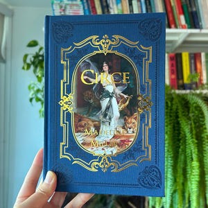 May include: A blue hardcover book titled Circe by Madeline Miller, featuring a central illustration of a figure in a white dress surrounded by lions, framed by intricate gold foil detailing on the front cover with decorative corner patterns.