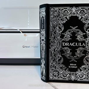 K&ouml;nnte beinhalten: Ein schwarzes Buch mit einem silbernen, gepr&auml;gten Einband, der ein kompliziertes Design und den Titel "Dracula" von Bram Stoker zeigt.