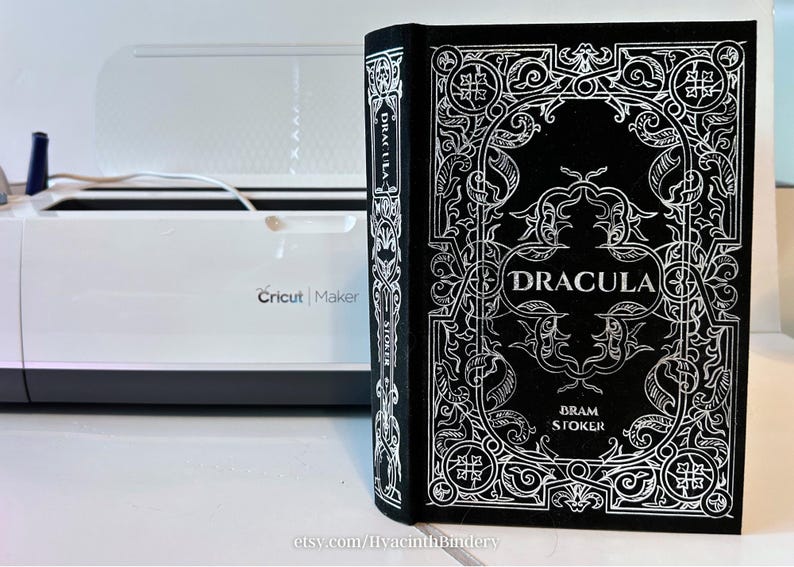 K&ouml;nnte beinhalten: Ein schwarzes Buch mit einem silbernen, gepr&auml;gten Einband, der ein kompliziertes Design und den Titel "Dracula" von Bram Stoker zeigt.