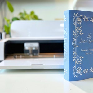 May include: A blue hardcover book titled "The Secret Garden" by Frances Hodgson Burnett. The book cover features a white floral design.
