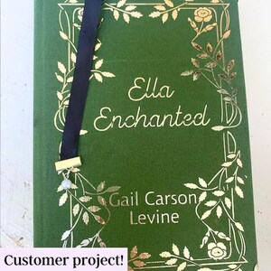 May include: A green book with gold leaf detailing and the title "Ella Enchanted" by Gail Carson Levine. A black ribbon bookmark with a gold charm and a pearl hangs from the book.