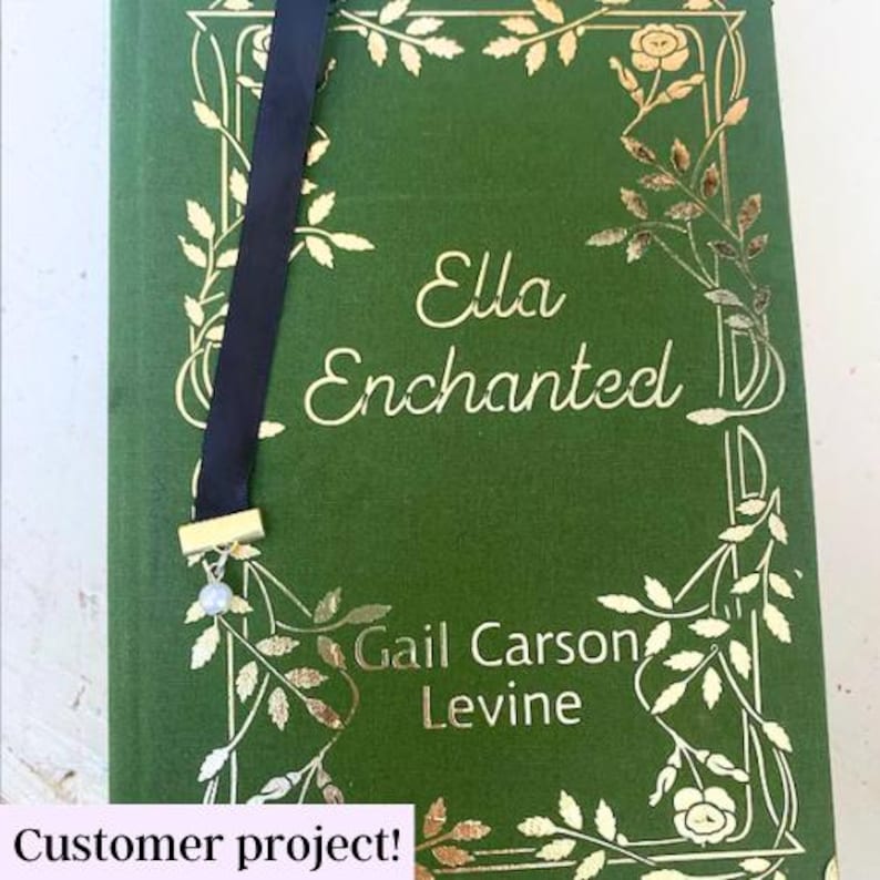 May include: A green book with gold leaf detailing and the title "Ella Enchanted" by Gail Carson Levine. A black ribbon bookmark with a gold charm and a pearl hangs from the book.
