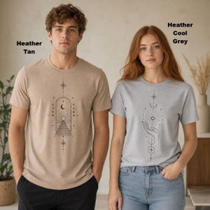 May include: A heather tan t-shirt with a black line art graphic of an arched doorway, stairs, and celestial symbols, and a heather cool grey t-shirt with a black line art graphic of a hand holding a sun, moon, and stars.