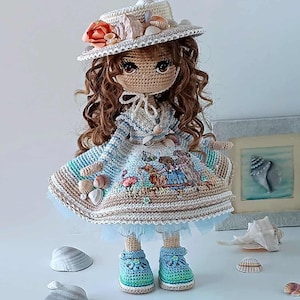May include: A handmade crochet doll wearing a white hat adorned with a coral-colored flower and seashells. The doll has brown curly hair, a blue and white dress, and blue and green shoes. The dress features a beach scene with seashells.