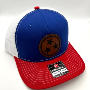 May include: A blue, white and red trucker hat with a brown leather patch featuring three white stars. The patch is centred on the front of the hat. The hat is adjustable and has the text "Richardson 112 Original Trucker Adjustable" on a white tag.