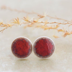 Può includere: Un paio di orecchini a bottone in ceramica rossa e bianca. Gli orecchini sono rotondi e hanno un bordo bianco con un centro rosso.