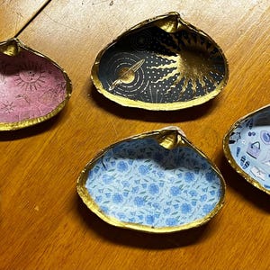 May include: A collection of decorative seashell dishes with gold-painted rims. The interiors feature various patterned designs, including floral, striped, and celestial motifs. The shells are arranged on a wooden surface.