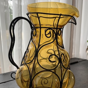 May include: A amber glass pitcher with a black metal cage design. The pitcher has a round body and a spout. The cage is made of intricate swirls and circles.
