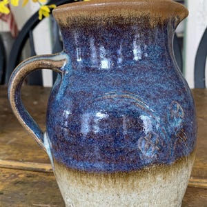 May include: Handmade ceramic pitcher with a blue and brown glaze. The pitcher has a handle and a spout. The bottom is a light beige color. The pitcher has a textured surface and is likely used for serving beverages.