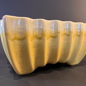 May include: A yellow ceramic planter with a rippled design. The planter has a light yellow glaze with a white glaze on the top edge.