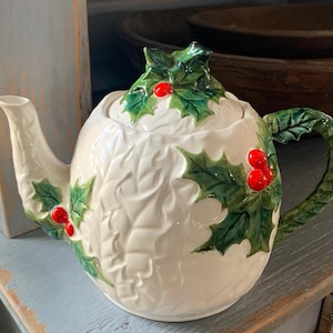 May include: A white ceramic teapot with a holly leaf and red berry design. The teapot has a curved spout and handle, and a lid with a holly leaf cluster. The holly leaves are green, and the berries are red. The teapot is on a light blue surface.