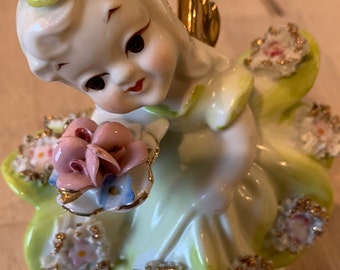 May include: A porcelain figurine of a girl with a pink flower crown and a green dress. She is holding a bouquet of pink and blue flowers. The figurine is decorated with gold accents and has a green base.