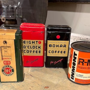 May include: A collection of vintage metal containers, including a green and yellow Sinclair HC Gasoline bank, two coffee tins labeled "Eight O'Clock" and "Bokar", and an orange and white "R-M Super-Max" can. The items are arranged on a wooden surface.