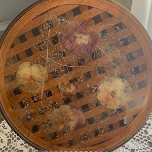 May include: Round wooden tray with a woven design, featuring dried flowers and delicate sprigs encased in clear resin. The tray has a dark brown border and a gold-colored metal edge.