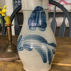 May include: A stoneware pitcher with a cream-colored base and blue floral designs. The pitcher has a wide body, a narrow neck, and a handle. The base of the pitcher has the text "Wisconsin Pottery, Columbus, WI 1989".