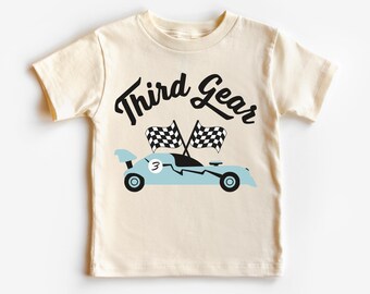 May include: A light beige t-shirt with the text "Third Gear" in black script. A blue race car with a checkered flag and the number "3" is printed below the text.