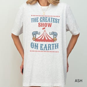 May include: A gray t-shirt with the text "The Greatest Show On Earth" printed on it. The text is surrounded by stars and there are two elephants balancing on balls under a red and white striped tent.
