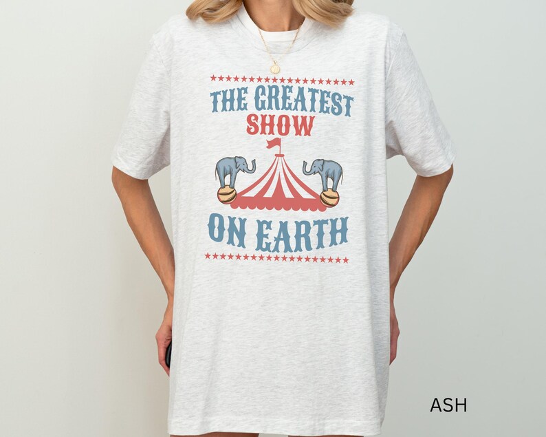 May include: A gray t-shirt with the text "The Greatest Show On Earth" printed on it. The text is surrounded by stars and there are two elephants balancing on balls under a red and white striped tent.