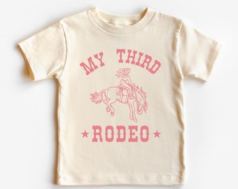 May include: A cream-colored t-shirt with a pink graphic of a cowgirl riding a horse. The text "MY THIRD RODEO" is printed in pink on the shirt.