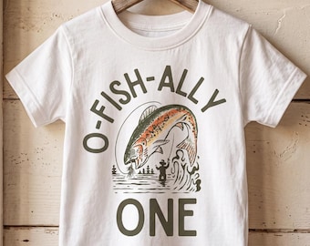 May include: A white short-sleeve t-shirt featuring a graphic of a jumping trout and a person fishing in a lake. The text O-FISH-ALLY is printed in dark green above the fish, and the word ONE is printed in dark green below the graphic.