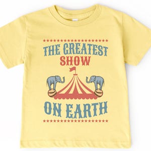 May include: A yellow t-shirt with a vintage circus theme. The text on the shirt reads "The Greatest Show On Earth" with a red and white striped tent and two elephants balancing balls.