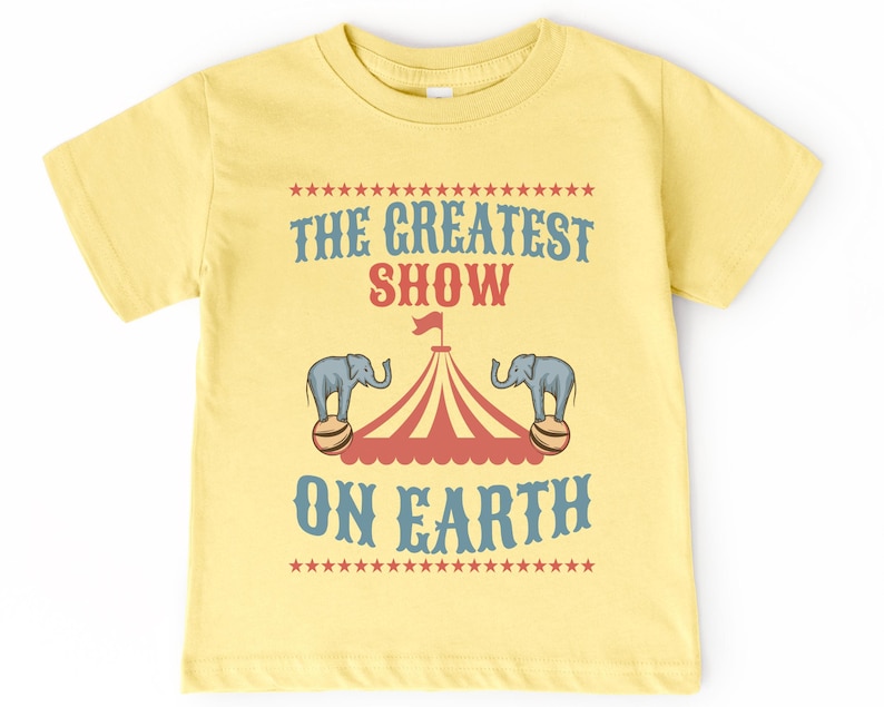May include: A yellow t-shirt with a vintage circus theme. The text on the shirt reads "The Greatest Show On Earth" with a red and white striped tent and two elephants balancing balls.
