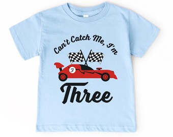 May include: Light blue toddler t-shirt with a red race car graphic and the text "Can't Catch Me, I'm Three." The race car has two checkered flags and the number 3. A fun shirt for a child's birthday.
